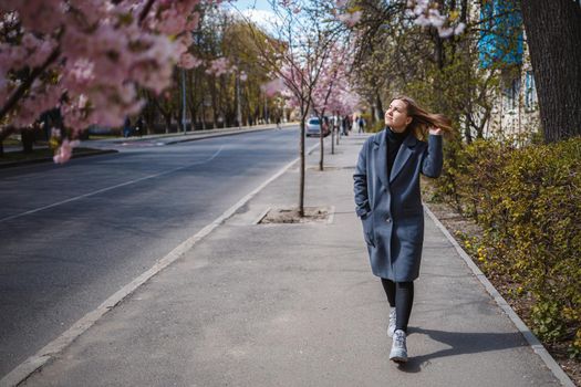 Sakura branches with flowers on a tree on the city streets. Happy woman girl in a gray palette walks along an alley with blooming sakura. Gorgeous fancy girl outdoors. Sakura tree blooming.