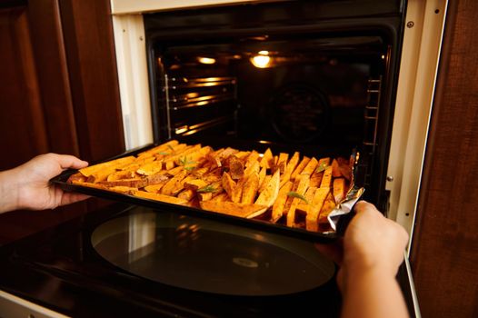 Close-up of housewife's hands putting a baking sheet with sliced sweet potatoes, seasoned with fragrant culinary herbs and spices and sprinkled with olive oil, into an oven while cooking vegan dinner