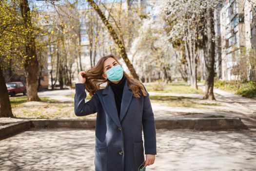 Girl, young woman in a protective sterile medical mask on her face, looking at the camera outdoors, in the spring garden. Air pollution, virus, pandemic coronavirus concept.