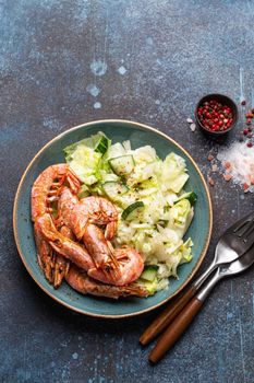 Whole fried big shrimps with green vegetables salad in blue bowl on stone rustic background from above. Fresh cooked delicious grilled shrimps served with veggies top view, healthy seafood meal