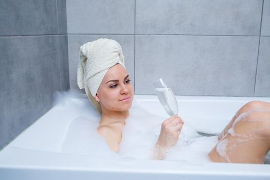 Girl woman with a white towel on her head lies with a glass of champagne in a white bathtub. In her hands a lot of pee soap. Full whirlpool foam. Relax after a hard day. Spa relaxing procedure