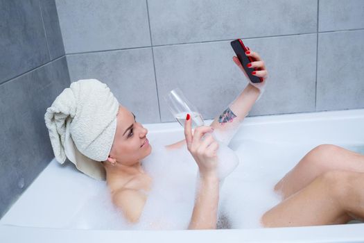 Girl woman with a towel on her head lies with a glass of champagne in a white bathtub and takes a selfie on the phone. In her hands a lot of drunk soap. Relax after a hard day. Spa relaxing procedure