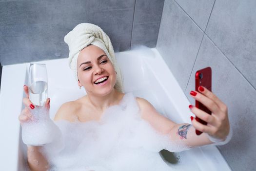 Girl woman with a towel on her head lies with a glass of champagne in a white bathtub and takes a selfie on the phone. In her hands a lot of drunk soap. Relax after a hard day. Spa relaxing procedure