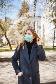 Girl, young woman in a protective sterile medical mask on her face, looking at the camera outdoors, in the spring garden. Air pollution, virus, pandemic coronavirus concept.
