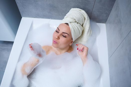 Girl woman with a white towel on her head lies in the bathroom in a white bath. In the hands of a lot of muddy soap. Full whirlpool foam. Relax after a hard day. Spa relaxing treatment