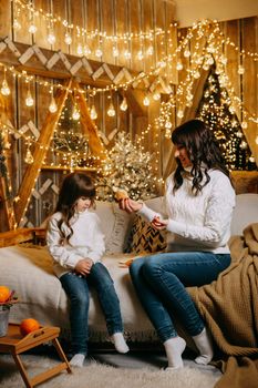 A little girl with her mother in a cozy home environment on the sofa next to the Christmas tree. The theme of New Year holidays and festive interior with garlands and light bulbs,