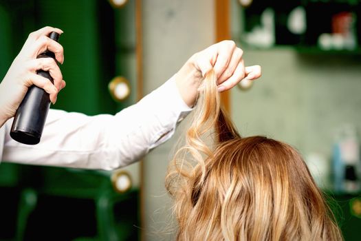 Female coiffeur fixing hairstyling of blonde woman with hairspray in a beauty salon