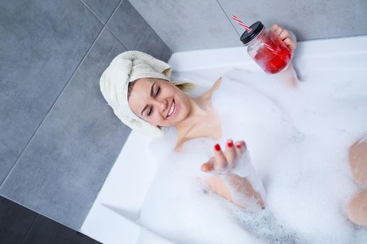 Beautiful young woman with a towel on her head, drinking a cocktail, takes a bath at home. Relax after a hard day.