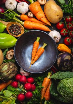 Two fresh farm carrots in vintage bowl and assorted organic vegetables on rustic black concrete background. Autumn harvest, vegetarian food or cooking clean healthy meal concept, top view