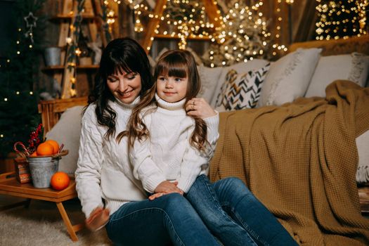 A little girl with her mother in a cozy home environment on the sofa next to the Christmas tree. The theme of New Year holidays and festive interior with garlands and light bulbs