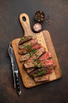 Grilled or fried prime marbled meat steak Ribeye sliced and served on wooden cutting board with cutlery. Top view of juicy cooked beef steak cut in slices on rustic brown concrete background