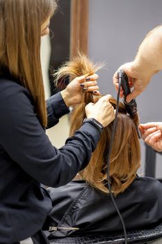 The rear view of two hairdressers are curling hair for a young woman with electric hair iron in a beauty salon