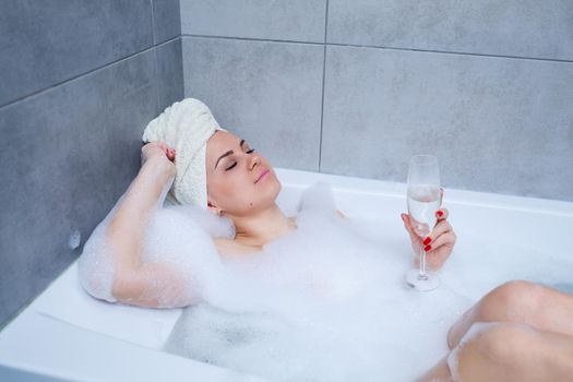Young beautiful woman with a white towel on her head lies in a white bath. In the hands of a glass of white wine. Relax after a hard day. Relaxing spa treatments