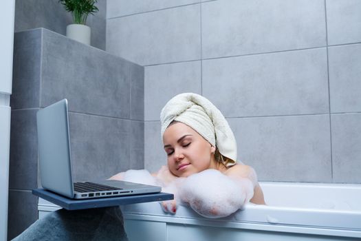 A young woman with a towel on her head is watching a movie on a laptop while sitting in a bath in a beauty salon. Relax in the bathroom without linen. Body care and relaxation concept.