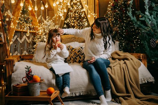 A little girl with her mother in a cozy home environment on the sofa next to the Christmas tree. The theme of New Year holidays and festive interior with garlands and light bulbs