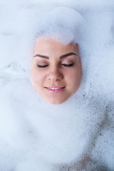 Closeup portrait of a girl in a bathtub and white foam around her face. Different emotions, grimaces, woman smile