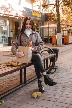 A stylish brunette woman walks around the autumn city. The brunette is sitting on a bench outside with a glass of coffee and a phone, holding a craft bag with a baguette.