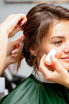 Makeup artist applying cream blush foundation tube on the cheek of the young caucasian woman in a beauty salon