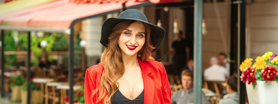 Smiling portrait of fashion young caucasian woman is posing in black hat and red jacket in the city street