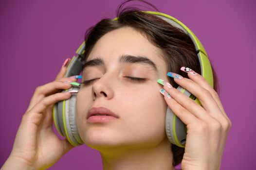 Sensual studio portrait of young pretty girl with short haircut and extravagant nail art listening to music in headphones. Eyes closed, colored background