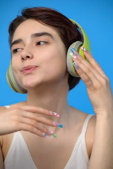 Portrait of cute young bare shoulders brunette with extravagant nail art listening to music using wireless headphones, studio shot