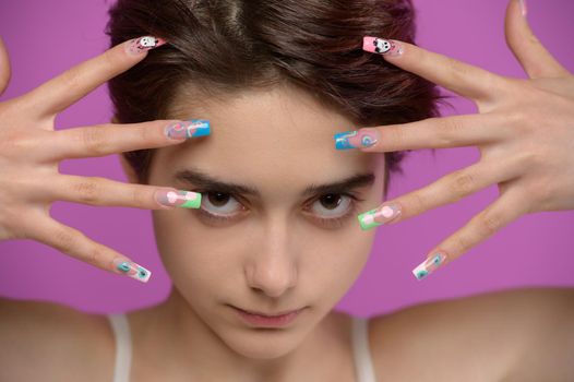 Pretty girl with short haircut and extravagant nail art looking into camera, studio portrait at colored background