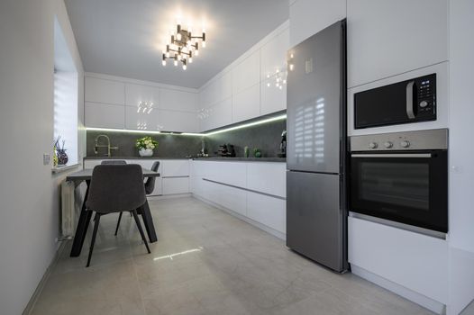 Luxurious modern trendy white and grey kitchen interior after renovation, with granite counter top and marble floor, dining table next to window