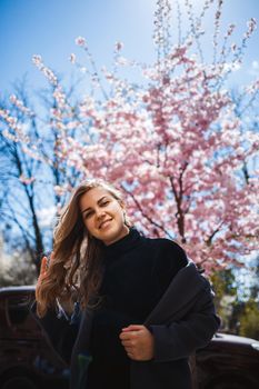 Sakura branches with flowers on a tree on the city streets. Stylish woman girl stands in the street with blooming sakura. Modern fashionable girl outdoors. Sakura tree blossoms.