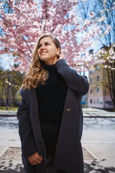 A young slender female model with long wavy hair and, dressed in a gray coat, sneakers, stands on the street near a flowering shrub with beautiful pink flowers in the background and poses.