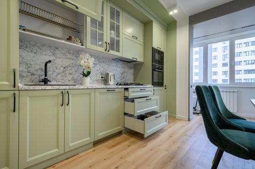 Modern trendy light green luxurious kitchen with dining table and dark green chairs, some drawers are retracted