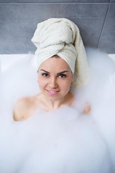 Girl woman with a white towel on her head lies in the bathroom in a white bath. Foam for around. Relax after a hard day. Spa relaxing treatment