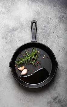 Black cast iron frying pan with ingredients for cooking: oil, fresh green rosemary, raw garlic cloves and colorful peppercorns on gray stone background flat lay top view, cooking food concept