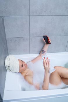 Girl woman with a towel on her head lies with a glass of champagne in a white bathtub and takes a selfie on the phone. In her hands a lot of drunk soap. Relax after a hard day. Spa relaxing procedure