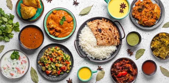 Indian ethnic food buffet on white concrete table from above: curry, samosa, rice biryani, dal, paneer, chapatti, naan, chicken tikka masala, mango lassi, dishes of India for dinner background