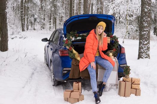 A woman in a winter snow-covered forest in the trunk of a blue car decorated with Christmas decor. The concept of Christmas and winter holidays.