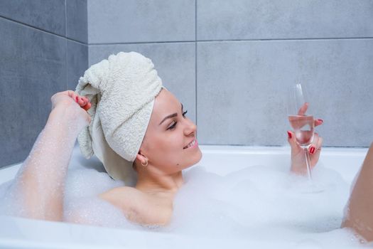 Girl woman with a white towel on her head lies with a glass of champagne in a white bathtub. In her hands a lot of pee soap. Full whirlpool foam. Relax after a hard day. Spa relaxing procedure