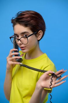 Beautiful young skinny brunette in yellow T-shirt talking quietly on a landline phone, studio shot at blue background