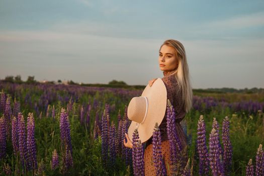 A beautiful woman in a straw hat walks in a field with purple flowers. A walk in nature in the lupin field.