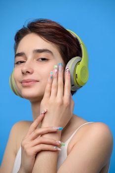 Portrait of cute young bare shoulders brunette with extravagant nail art listening to music using wireless headphones, studio shot