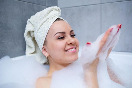 Girl woman with a white towel on her head lies in the bathroom in a white bath. In the hands of a lot of muddy soap. Full whirlpool foam. Relax after a hard day. Spa relaxing treatment