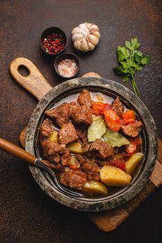 Delicious beef meat stew dish with potatoes, carrot and gravy in rustic metal bowl with spoon, bunch of fresh parsley, garlic cloves, spices on brown concrete background top view