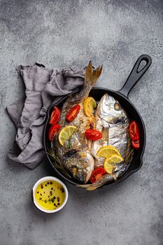 Roasted or fried fresh fish sea bream or dorado with lemon, herbs and tomatoes in cast iron skillet great for healthy meal or Mediterranean diet on rustic grey stone background from above