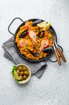Classic dish of Spain, seafood paella in traditional pan on white wooden background top view. Spanish paella with shrimps, clamps, mussels, green peas and fresh lemon wedges from above .