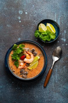 Traditional spicy Thai soup Tom Yum with shrimp and seafood in ceramic bowl on blue stone rustic background from above, classic dish of cuisine of Thailand