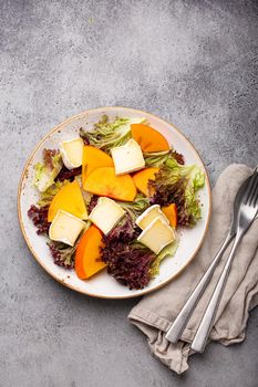 Top view flat lay persimmon salad with brie cheese and fresh salad leaves on white plate and stone gray background, seasonal fruit salad as appetizer, vegetarian healthy food