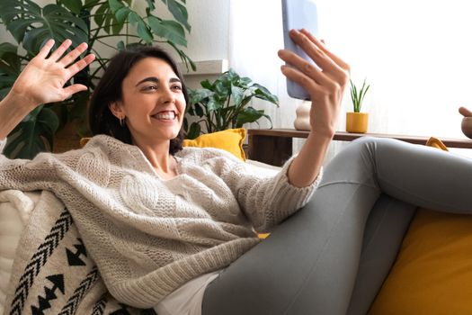 Young smiling caucasian woman relaxing at home waves hand during cellphone video call. Technology concept.
