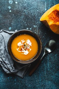 Homemade pumpkin cream soup served in brown ceramic bowl on dark stone background with spoon decorated with cut fresh pumpkin, top view. Traditional autumn comfort food.