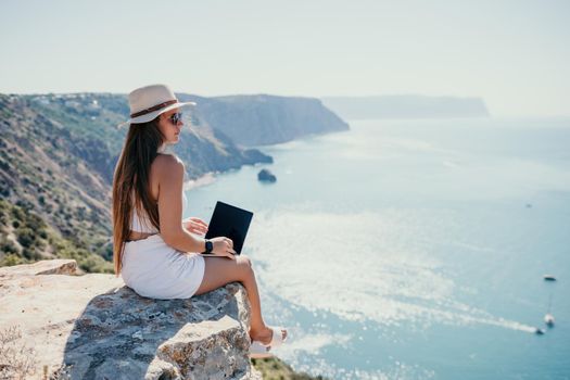 Successful business woman in yellow hat working on laptop by the sea. Pretty lady typing on computer at summer day outdoors. Freelance, travel and holidays concept.