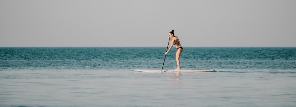 Silhouette of woman standing, surfing on SUP board, confident paddling through water surface. Idyllic sunset or sunrise. Sports active lifestyle at sea or river.