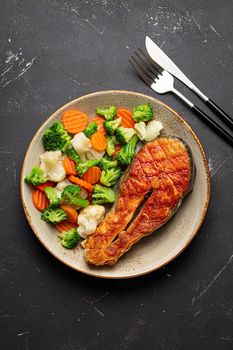 Healthy fish dinner: grilled salmon fish steak with vegetables salad on ceramic plate with fork and knife on black slate stone background from above, healthy clean and diet eating concept